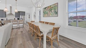 Une cuisine moderne dotée d’un îlot central, d’un coin repas avec une longue table et des chaises, et d’une grande fenêtre avec des stores.