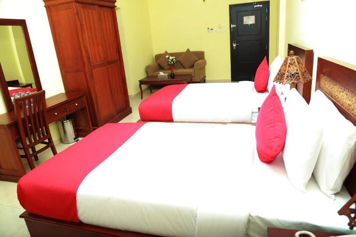 Two beds with red and white bedding, a wooden wardrobe, a small table with a vase, and a sofa in a room with yellow walls.