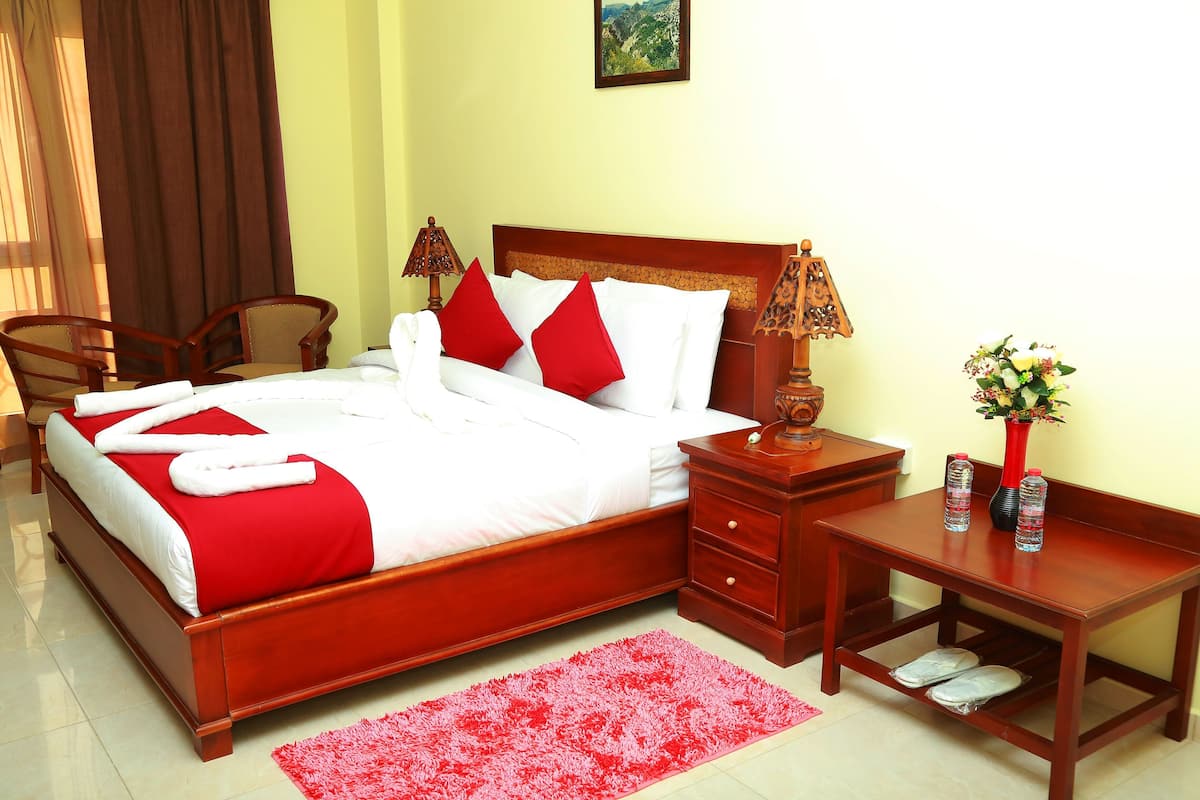 A hotel room with a wooden bed, red pillows, a red rug, a wooden nightstand, a chair, and a small table with a vase and bottles.