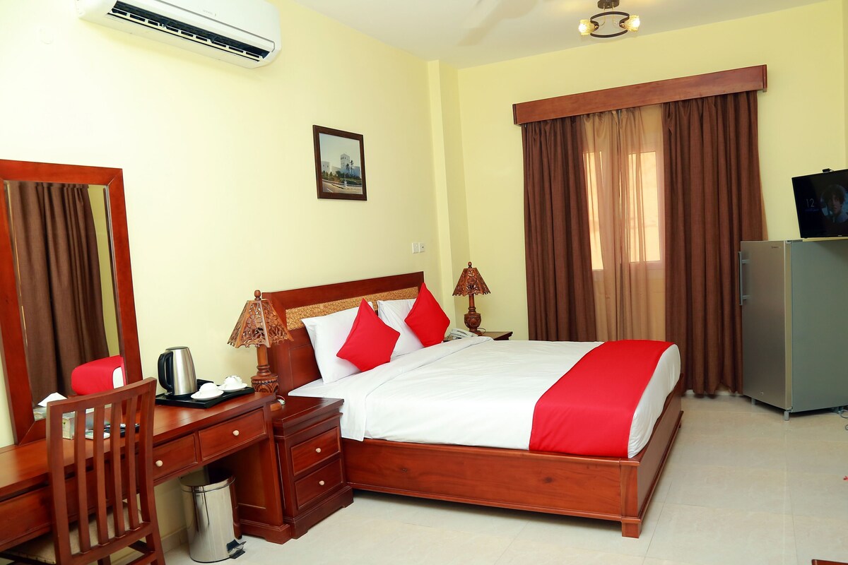 A hotel room with a wooden bed, a desk with a chair, a television, and a refrigerator.