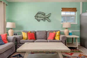 A living room with a teal wall, a fish wall decoration, a gray sofa with colorful cushions, a wooden coffee table, and a window with a bamboo blind. - Close to the beach! Water view, pet friendly! - Nautical Nonsense (Dauphin Island)