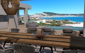 A terrace with a wooden table, chairs, and a bowl of fruit. A pool is visible in the background with a view of the sea and a hillside town.