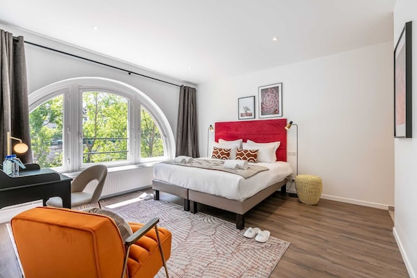Une chambre moderne avec une fenêtre incurvée, un lit doté d’une tête de lit rouge et un bureau avec une chaise.