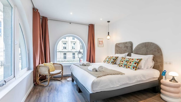 A modern bedroom with a large bed, a window with curtains, a wicker chair, and a view of buildings outside.