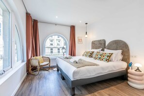 Une chambre moderne avec un grand lit, une fenêtre avec des rideaux, un fauteuil en osier et une vue sur les bâtiments environnants.