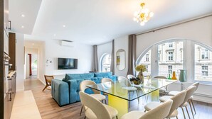 A modern living room with a glass dining table, blue sofa, and a TV mounted on the wall.