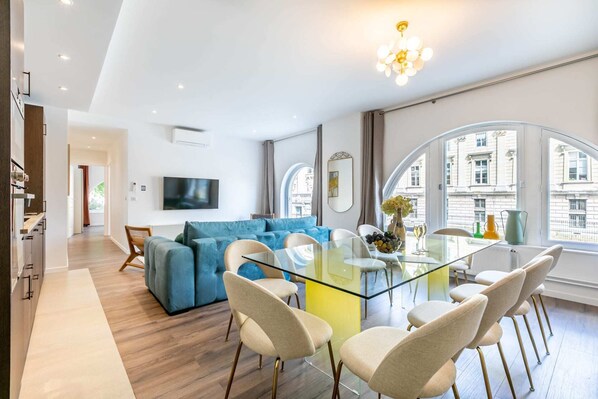 Un salon moderne avec une table à manger en verre, un canapé bleu et un téléviseur fixé au mur.
