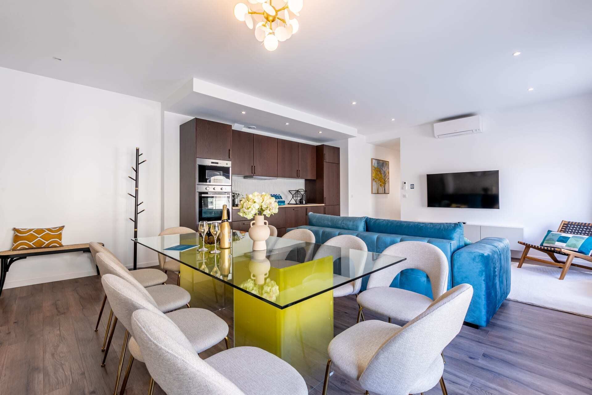 A modern living room with a glass dining table, a blue sofa, and a flat-screen TV mounted on the wall.