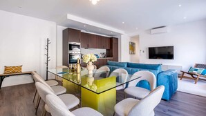 A modern living room with a glass dining table, a blue sofa, and a flat-screen TV mounted on the wall.