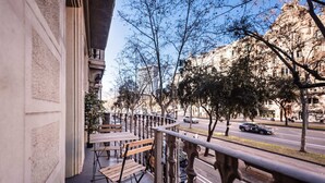 Un balcone con sedie di legno e ringhiera metallica che si affaccia su una strada con auto e alberi.