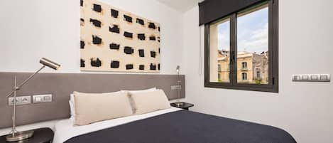 A bedroom with a large bed, a headboard, a window with a view of buildings, and a wall-mounted artwork.