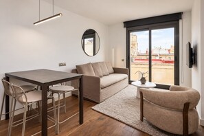 Een moderne woonkamer met een bank, een eettafel en stoelen, een klein bijzettafeltje met een vaas, en een groot raam met uitzicht op gebouwen.