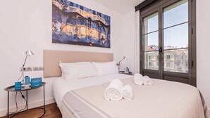 A hotel room with a large bed, a nightstand with a lamp, a painting of a building, and a window with a city view. - Sweett | Palau 602 - One Bedroom Apartment, Sleeps 3 (Barcelona)