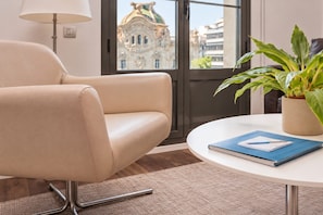 A modern room with a beige leather chair, a round table with a book, a potted plant, and a view of a historic building through a window. - Sweett | Palau 602 - One Bedroom Apartment, Sleeps 3 (Barcelona)