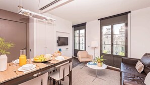 A modern living room with a dining area, a sofa, a TV, and a large window with a view of buildings. - Sweett | Palau 602 - One Bedroom Apartment, Sleeps 3 (Barcelona)