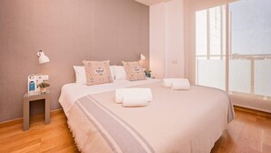 Un dormitorio con una cama grande, dos lámparas de noche, una mesita con una planta y una ventana con cortinas translúcidas.