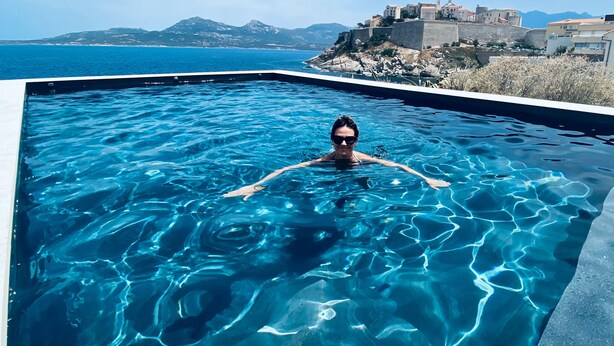 Eine Person schwimmt in einem klaren, blauen Pool mit Blick auf eine Küstenstadt und Berge im Hintergrund.