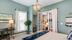 A bedroom with a bed, a desk, a chair, a chandelier, and a view into another room.