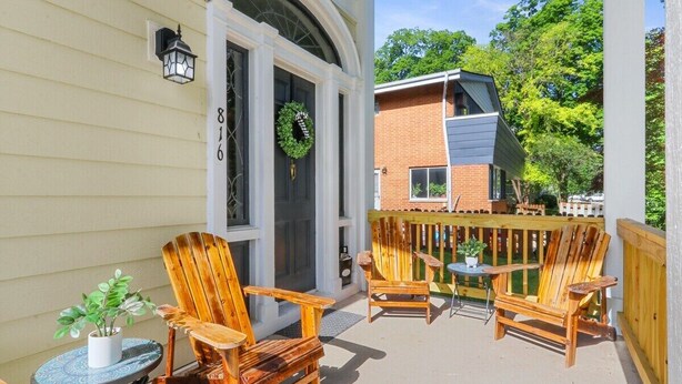 A covered patio with wooden chairs and a small table, a door with a wreath, and a brick building in the background.