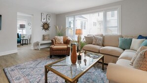 A living room with a sofa, armchairs, a coffee table, and a rug. There is a window with blinds, a lamp, and a decorative wall piece.