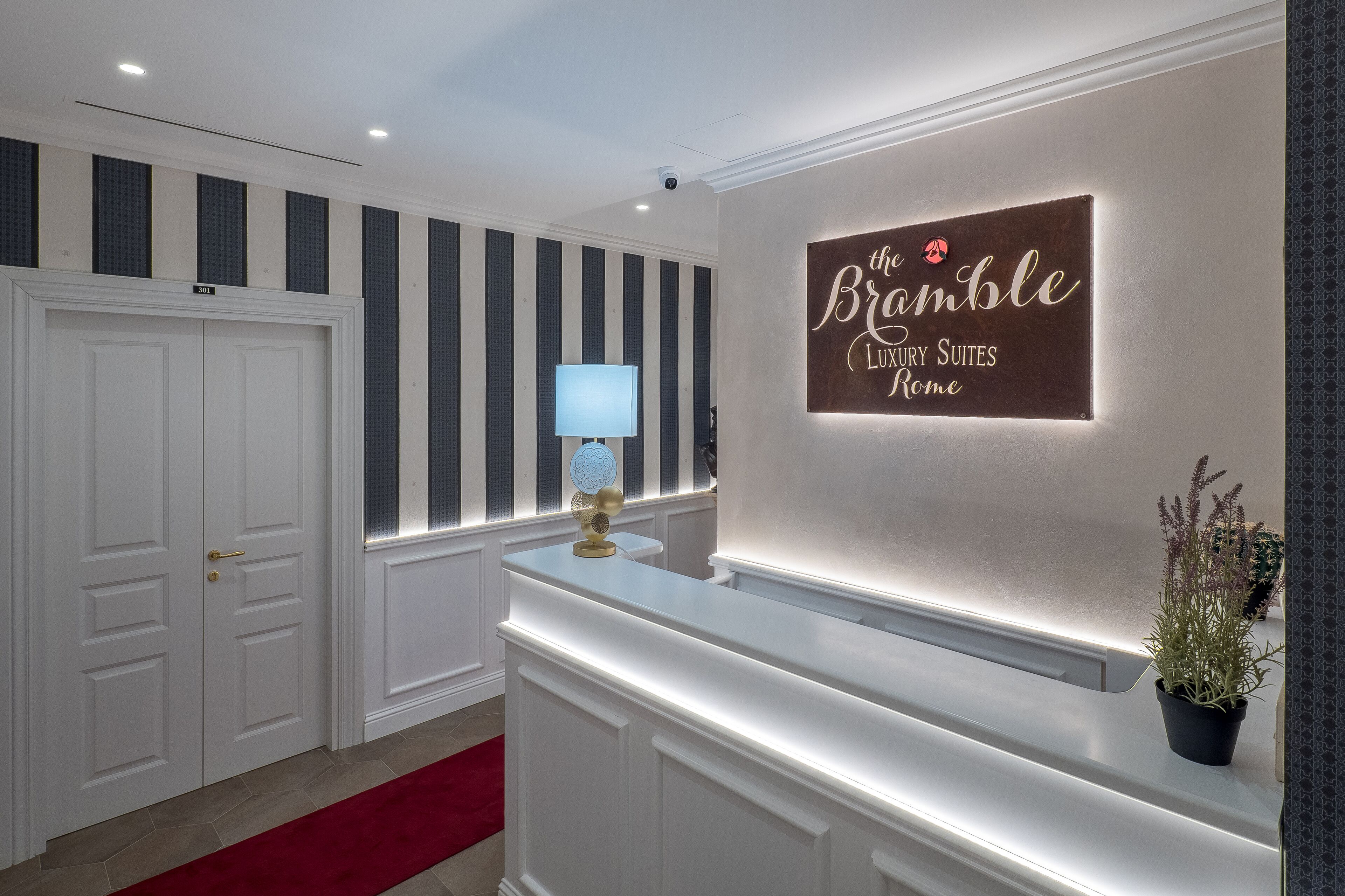 A reception area with a sign reading "The Bramble Luxury Suites Rome," a white reception desk, a potted plant, and a red carpet.