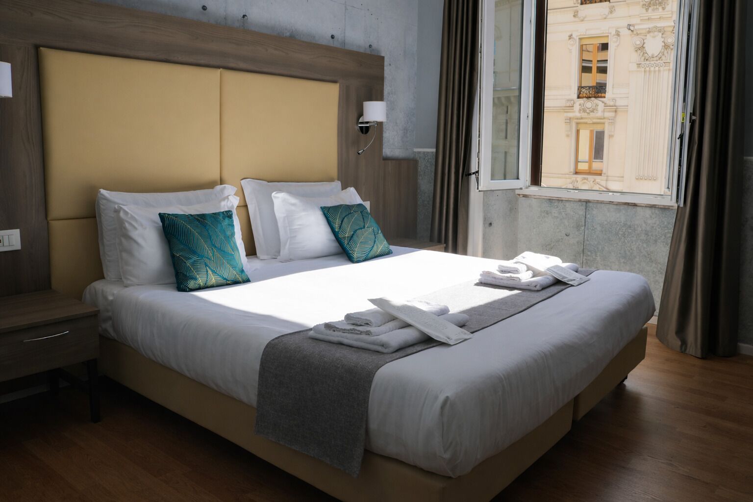 A hotel room with a large bed, a nightstand, a window with curtains, and a view of buildings outside.