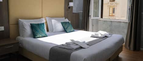 A hotel room with a large bed, a nightstand, a window with curtains, and a view of buildings outside.