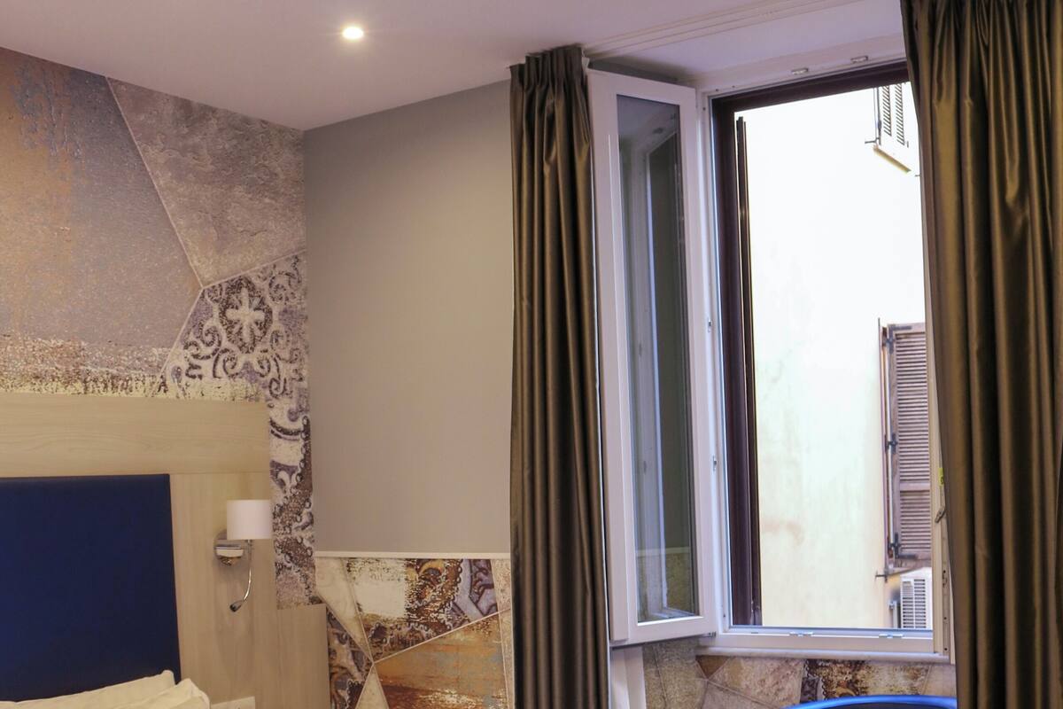 A hotel room with a bed, a chair, a window with curtains, and a wall with a patterned design.