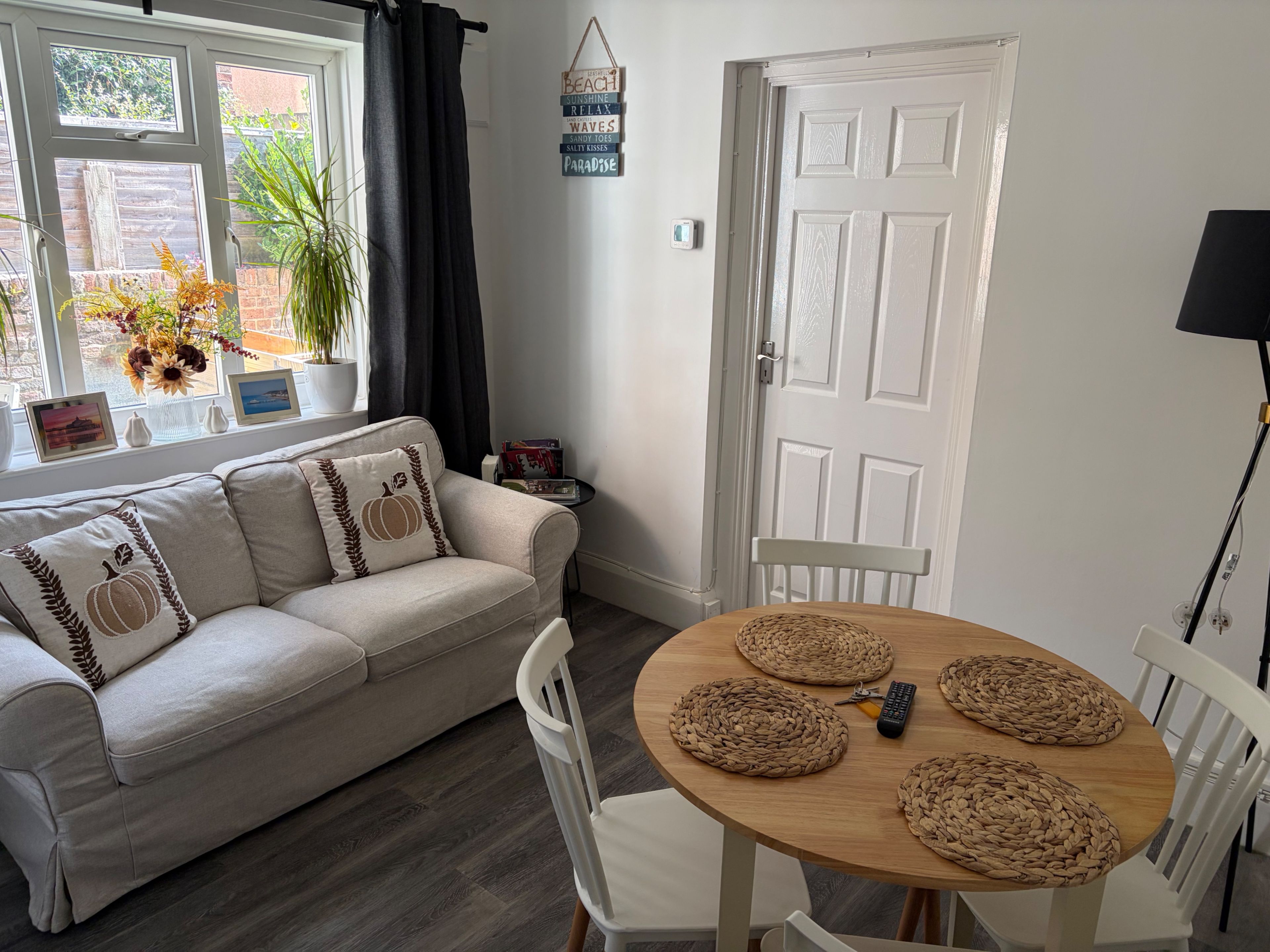 Ein gemütliches Wohnzimmer mit einer Couch, einem Holzesstisch mit Platzsets, einer weißen Tür und einem Fenster mit Vorhängen.