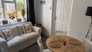Ein gemütliches Wohnzimmer mit einer Couch, einem Holzesstisch mit Platzsets, einer weißen Tür und einem Fenster mit Vorhängen.