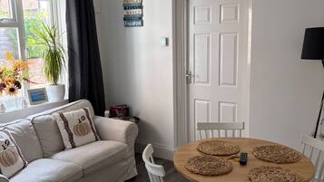 Una habitación con un sofá blanco, una mesa redonda de madera con sillas, una puerta y una ventana con cortinas.