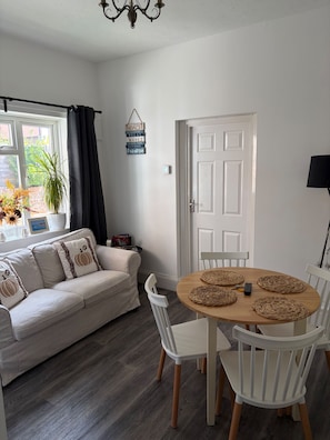A room with a white sofa, a round wooden table with chairs, a door, and a window with curtains. - 2-bed, Sleeps 4, Free Parking, Pets Allowed (Eastbourne)