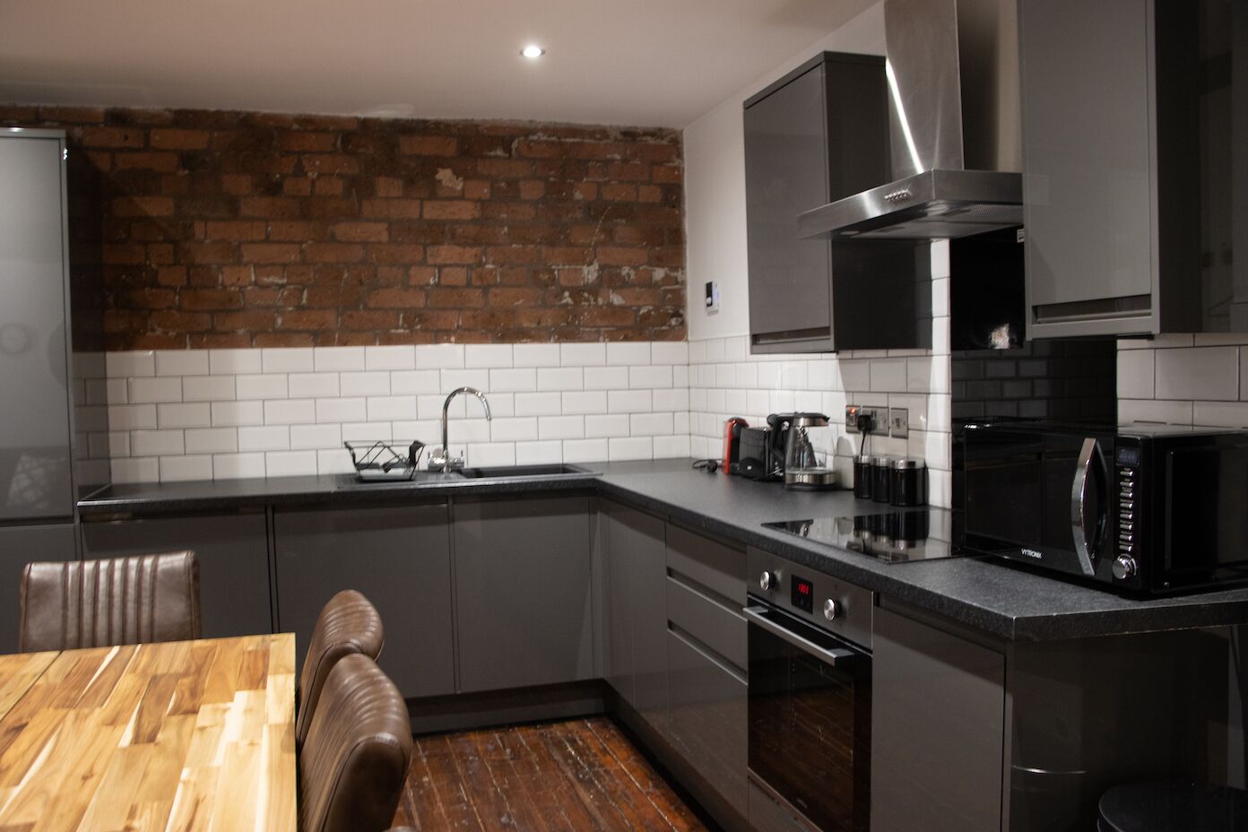 A modern kitchen with grey cabinets, a brick wall, a stainless steel range hood, a microwave, and a coffee maker.