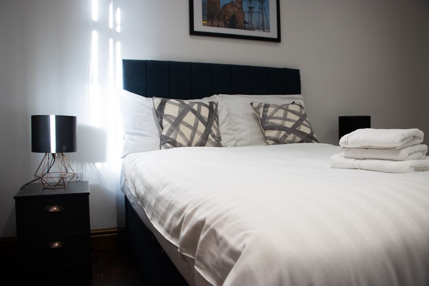 A neatly made bed with white linens, a dark headboard, and patterned pillows. There are two bedside tables with lamps and a folded towel on the right side.