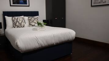 A neatly made bed with white linens and patterned pillows, a dark blue headboard, and a small plant on the bed.