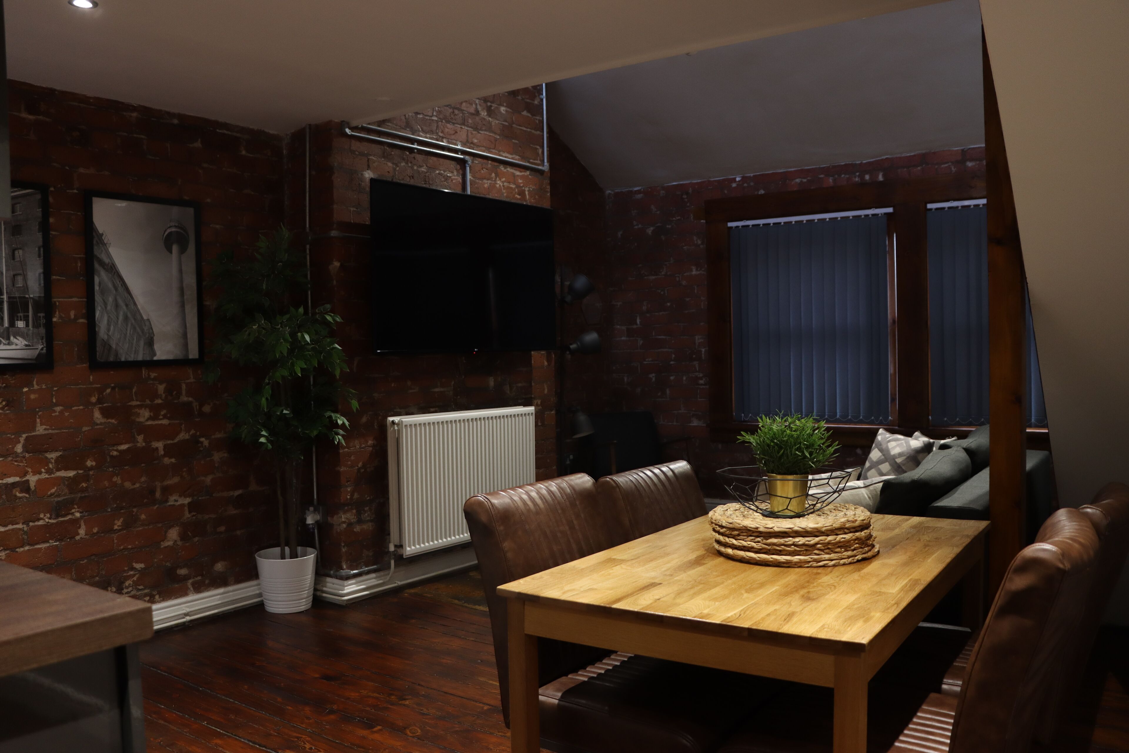 A dining area with a wooden table, leather chairs, a potted plant, and a brick wall.