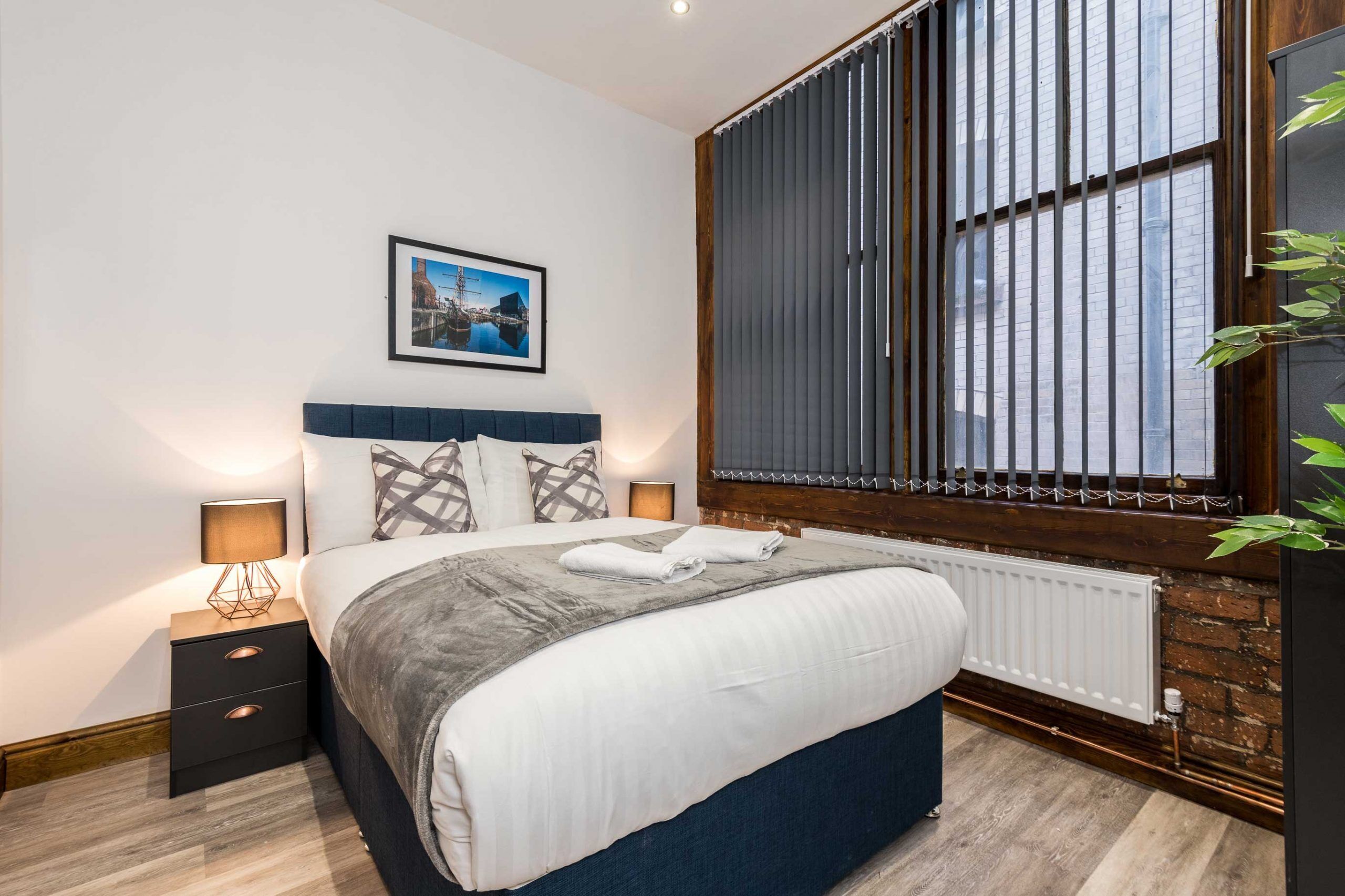 A modern bedroom with a large bed, a nightstand with a lamp, a framed picture on the wall, and a window with blinds.