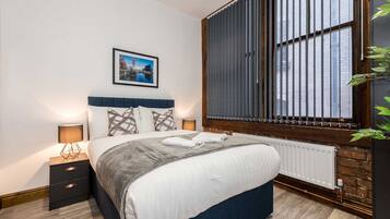A modern bedroom with a large bed, a nightstand with a lamp, a framed picture on the wall, and a window with blinds.