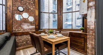 A cozy dining area with a wooden table set for two, a brick wall, and large windows offering a view of the city.