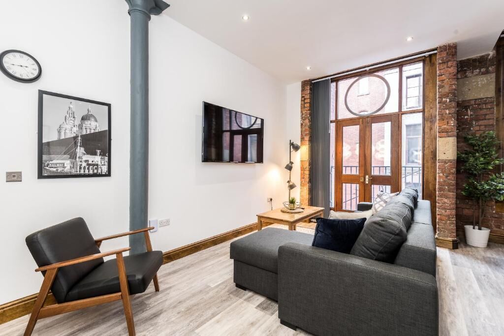 A modern living room with a grey sofa, a black chair, a wooden coffee table, and a large window with a view of a brick wall.