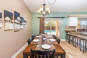 A dining area with a long wooden table set for six, surrounded by chairs, with a chandelier above.