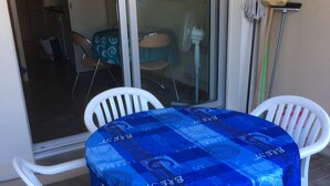 Un espace patio avec une table recouverte d’une nappe bleue, des chaises en plastique blanches et une porte vitrée donnant sur un espace intérieur.