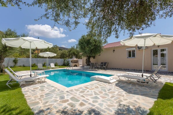 A swimming pool with stone tiles, surrounded by lounge chairs and umbrellas, and a building with a red roof in the background. - Michaelo Villa - Affordable Luxury - Michaelo Villa - Affordable Luxury (Zakynthos)