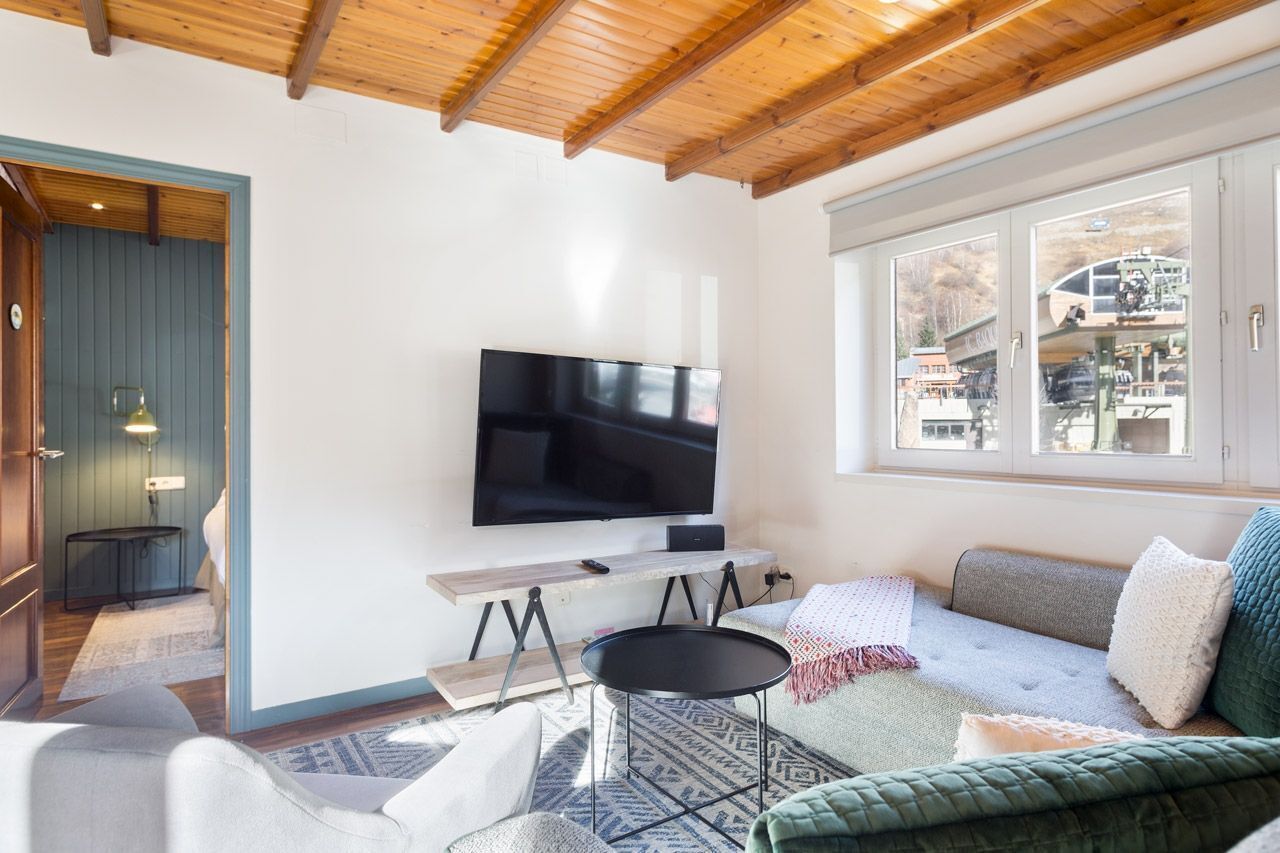 A modern living room with a flat-screen TV, wooden ceiling, and a large window offering a view of a building.