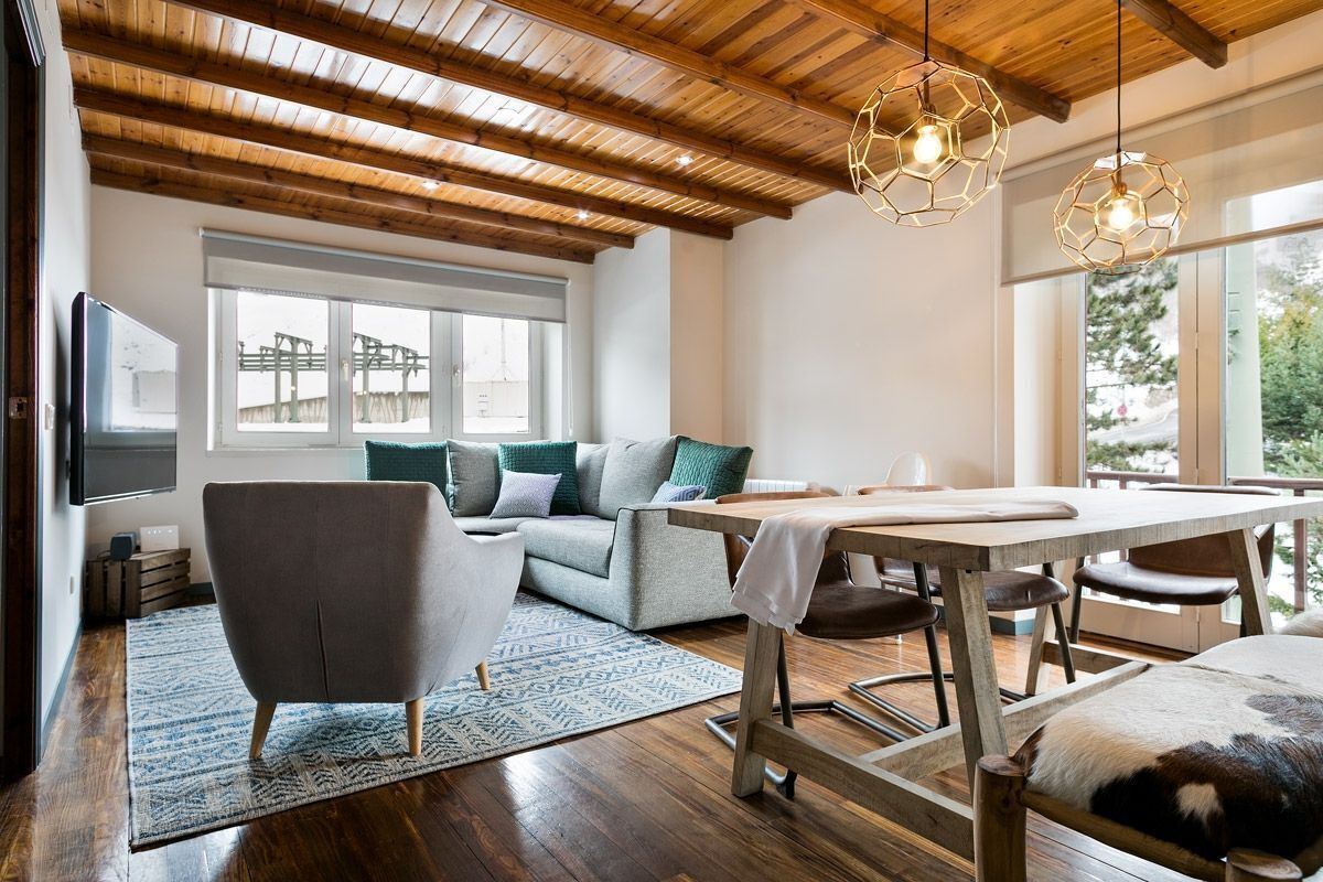 A modern living room with a wooden ceiling, a grey sofa with green cushions, a dining table with chairs, and a large window with a view of greenery.