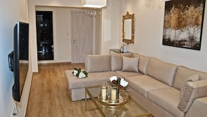 Un salon avec un canapé beige, une table basse en verre ornée de bougies, un téléviseur et un tableau représentant des arbres.