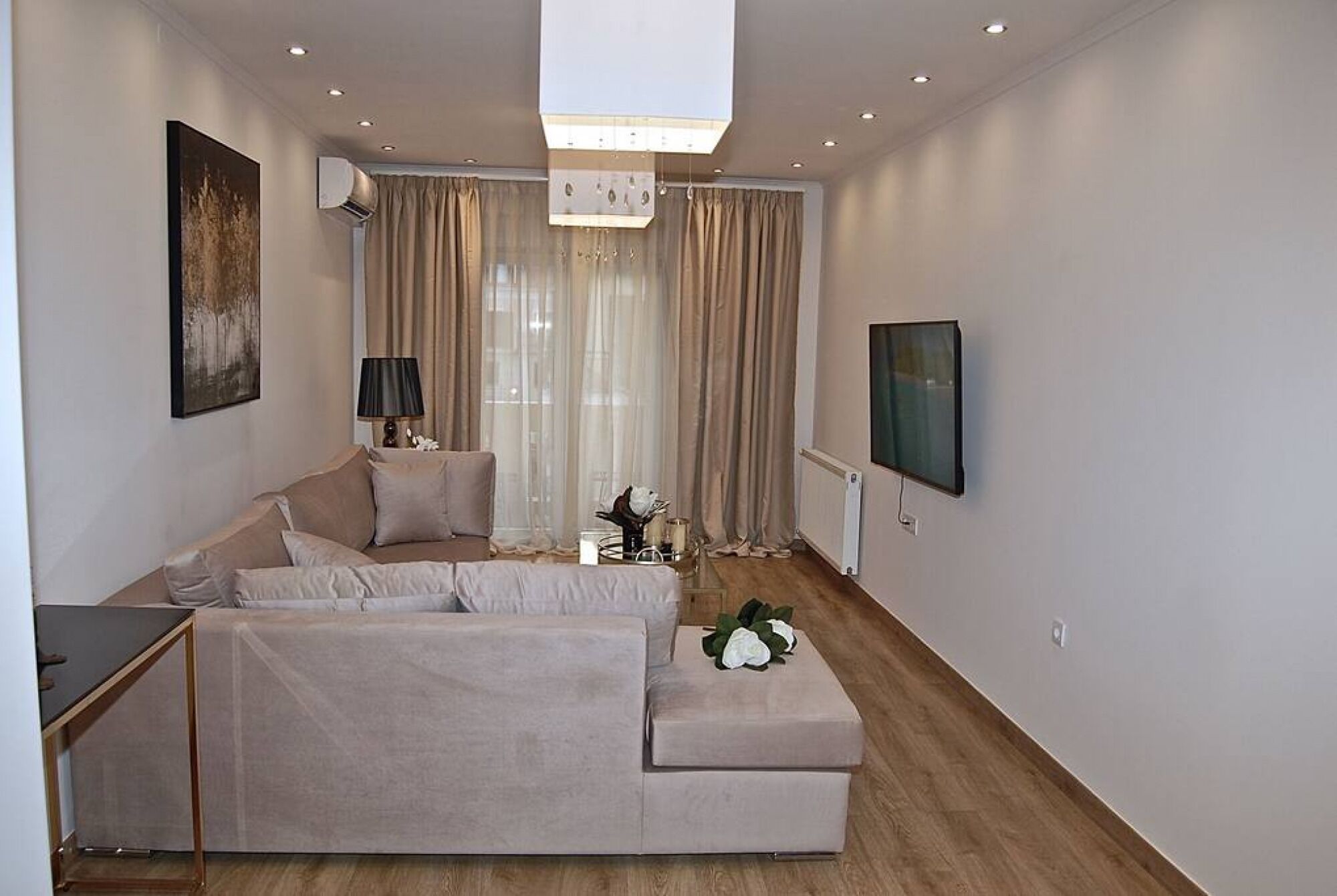 A modern living room with a large L-shaped sofa, a small table with a vase, and a flat-screen TV mounted on the wall.