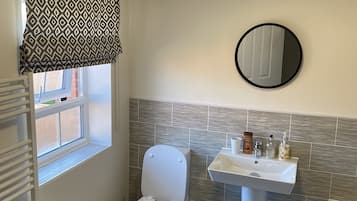 Une salle de bain avec des toilettes, un lavabo et un miroir. Il y a une fenêtre avec un store à motifs, un radiateur et une serviette violette sur le sol.