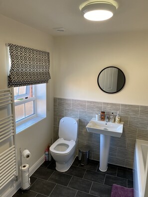 Une salle de bain avec des toilettes, un lavabo et un miroir. Il y a une fenêtre avec un store à motifs, un radiateur et une serviette violette sur le sol.