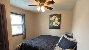 Une chambre à coucher comprenant un lit, un ventilateur de plafond, une fenêtre avec des stores et une œuvre d’art accrochée au mur.